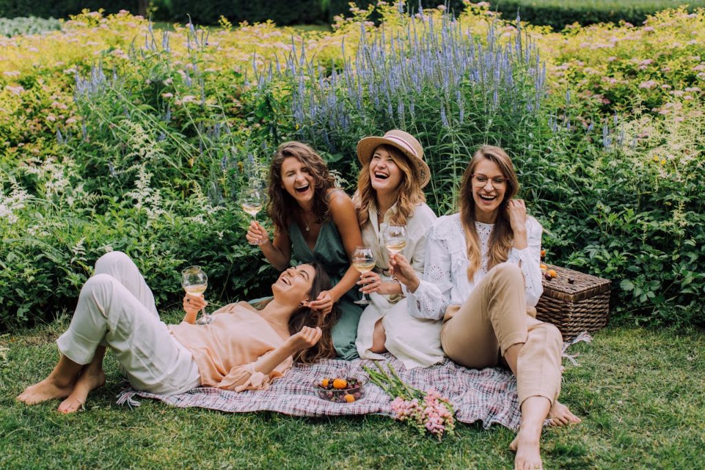 Four women enjoying a picnic, laughter, and wine in a vibrant summer garden.